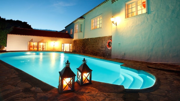 Un área de piscina con un deck de piedra, un edificio con techo de tejas rojas y dos faroles.