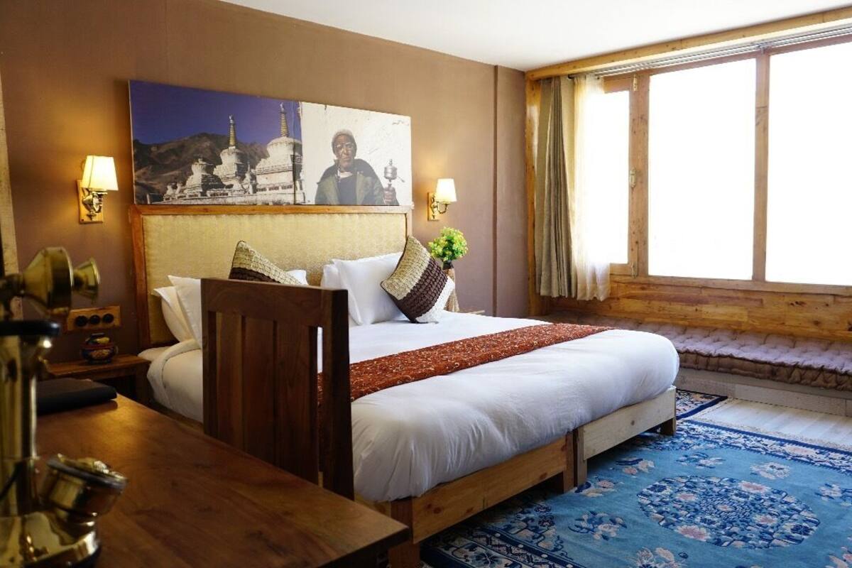 A hotel room with a large bed, a wooden headboard, a window with curtains, a bedside table with a lamp, and a wall-mounted picture.