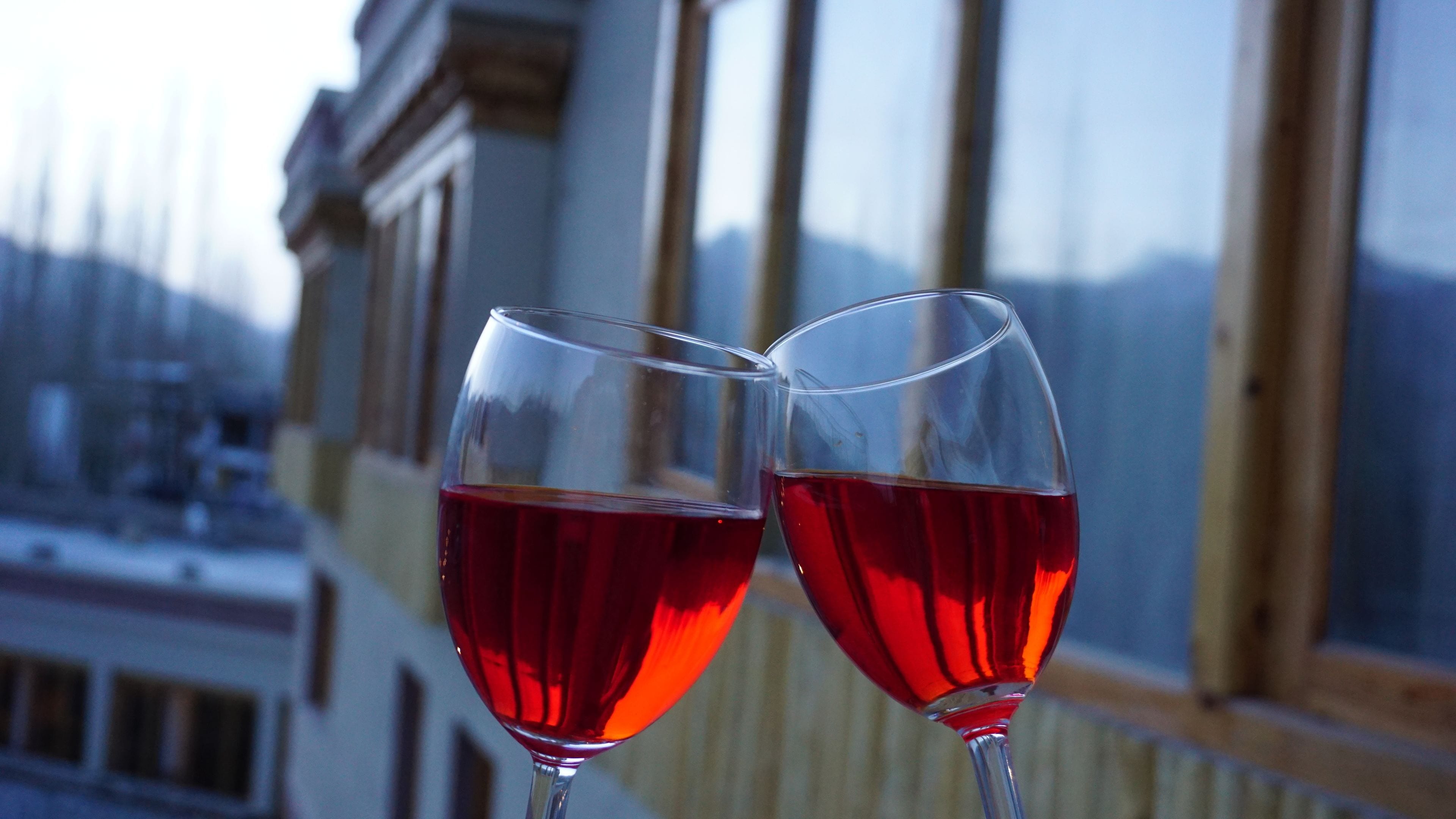 Two wine glasses filled with red wine on a balcony with a view of a building and a mountain in the background.