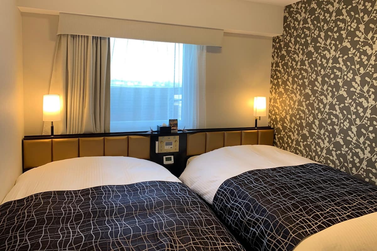 Habitació d'hotel amb dos llits, una finestra amb cortines, una taula de nit amb una làmpada i una paret amb un disseny floral.