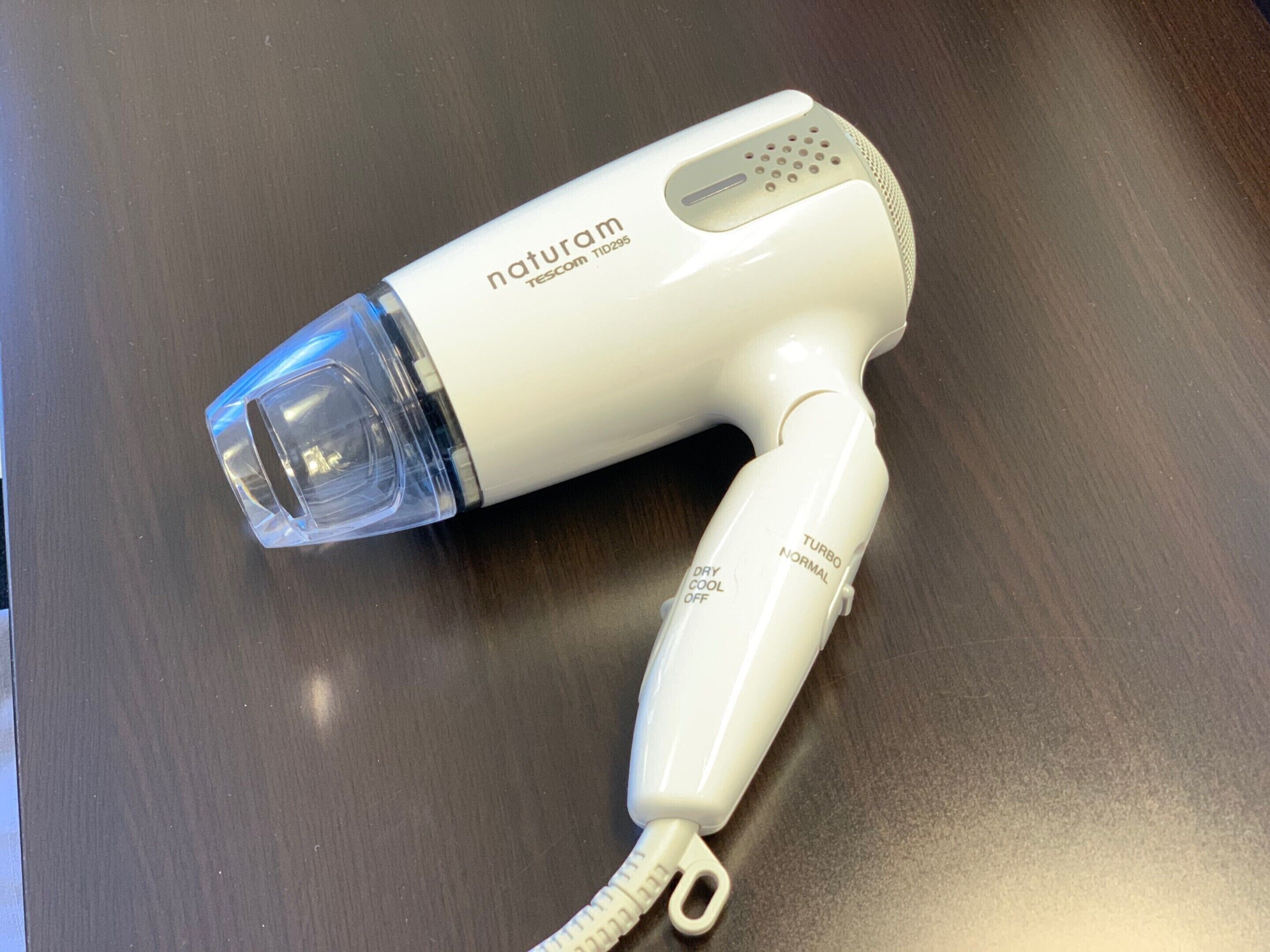 Un sèche-cheveux blanc, doté d’un embout transparent et d’un cordon, portant les inscriptions « naturam » et « TISCOM TID295 ».
