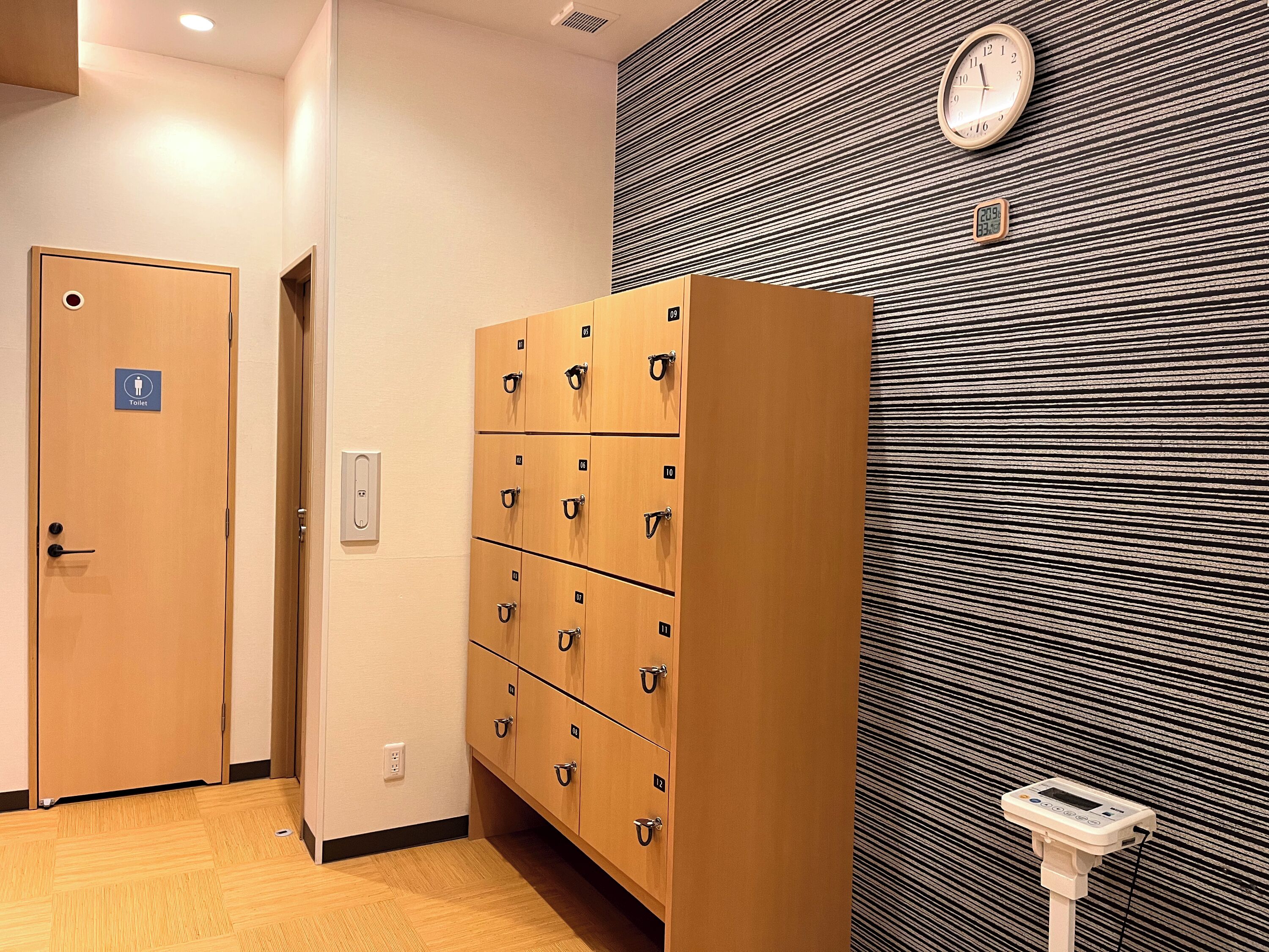 Habitación de hotel con una puerta de madera que indica "Toilet", varios taquillones de madera, un reloj en la pared y un enchufe.