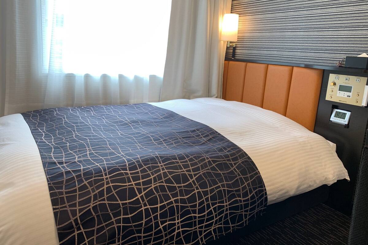 Habitació d'hotel amb llit, panell de control i finestra amb cortines.