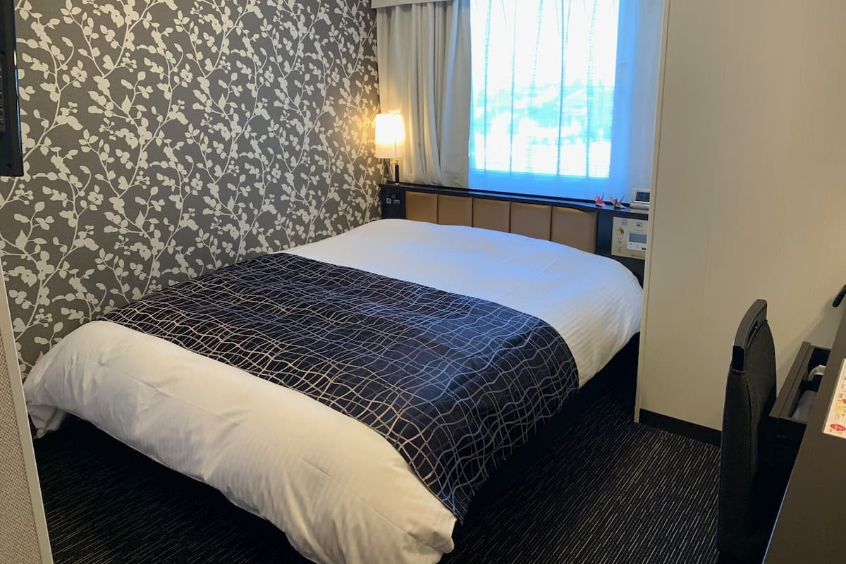 Habitació d'hotel amb un llit, una petita taula de nit, una làmpada i una finestra amb cortines.