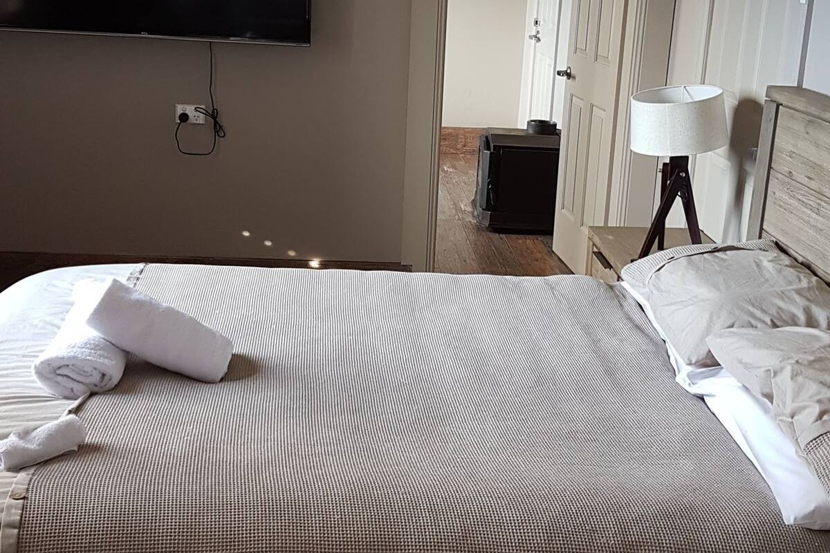 Habitación de hotel con cama, televisor de pantalla plana montado en la pared, mesita de noche con lámpara y puerta que da a otra habitación.