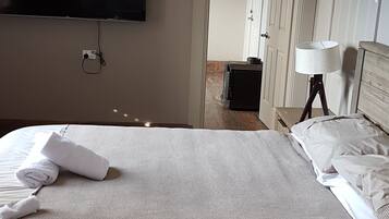 A hotel room with a bed, a flat-screen TV mounted on the wall, a bedside table with a lamp, and a door leading to another room.