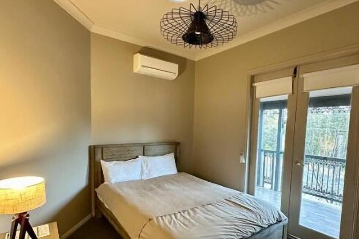 Un dormitorio con cama, ventilador de techo, equipo de aire acondicionado, mesita de noche y vistas al exterior a través de una puerta de vidrio.