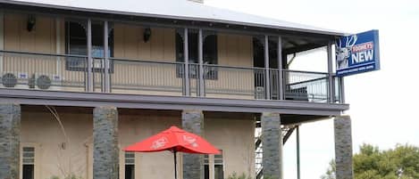 A two-story building with a balcony, stone pillars, and outdoor seating with red umbrellas.