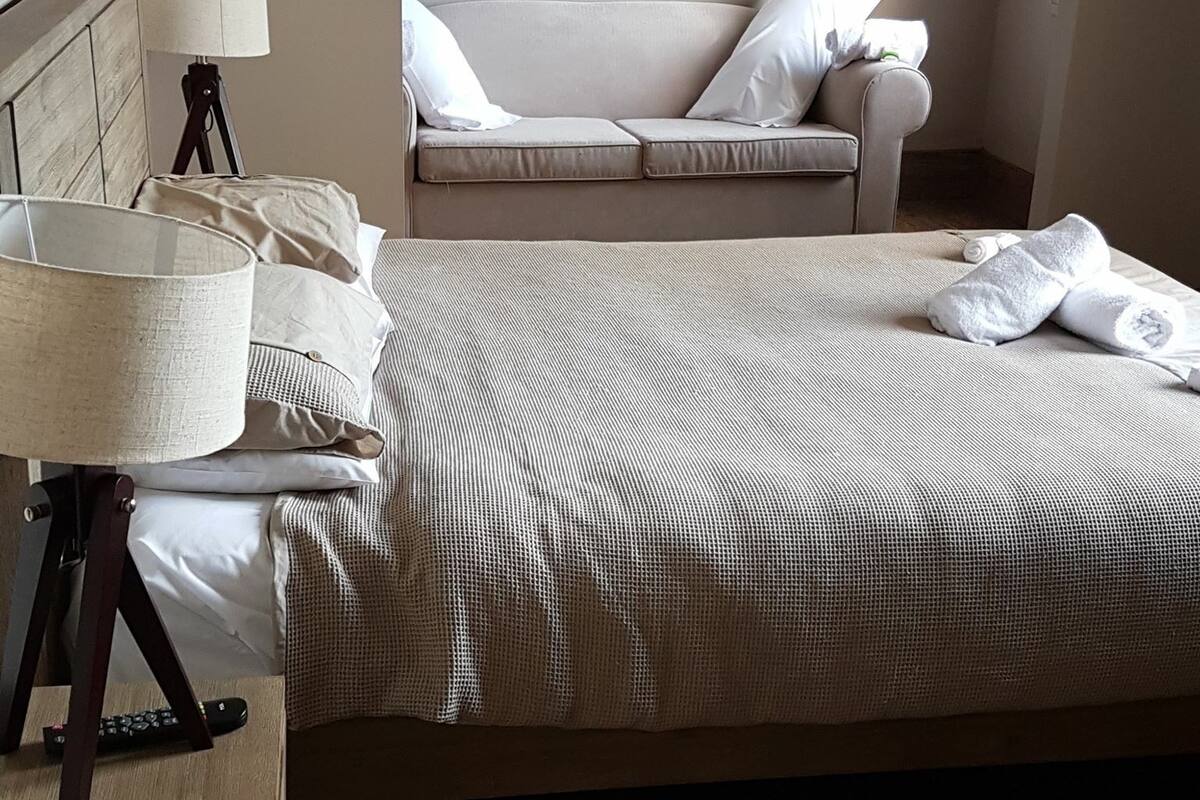Un dormitorio con cama, una lámpara de noche, un control remoto y un sofá al fondo.