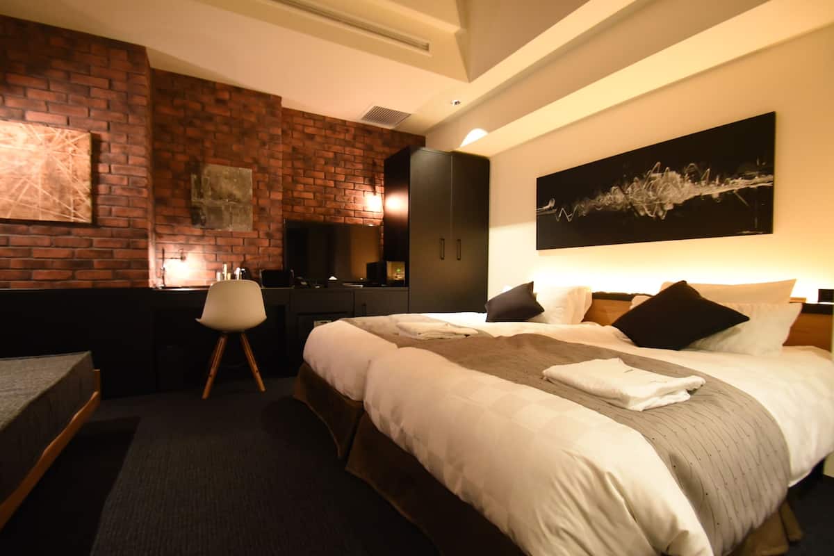 A hotel room with a brick accent wall, a large bed with white bedding, a desk with a chair, and a painting above the bed.