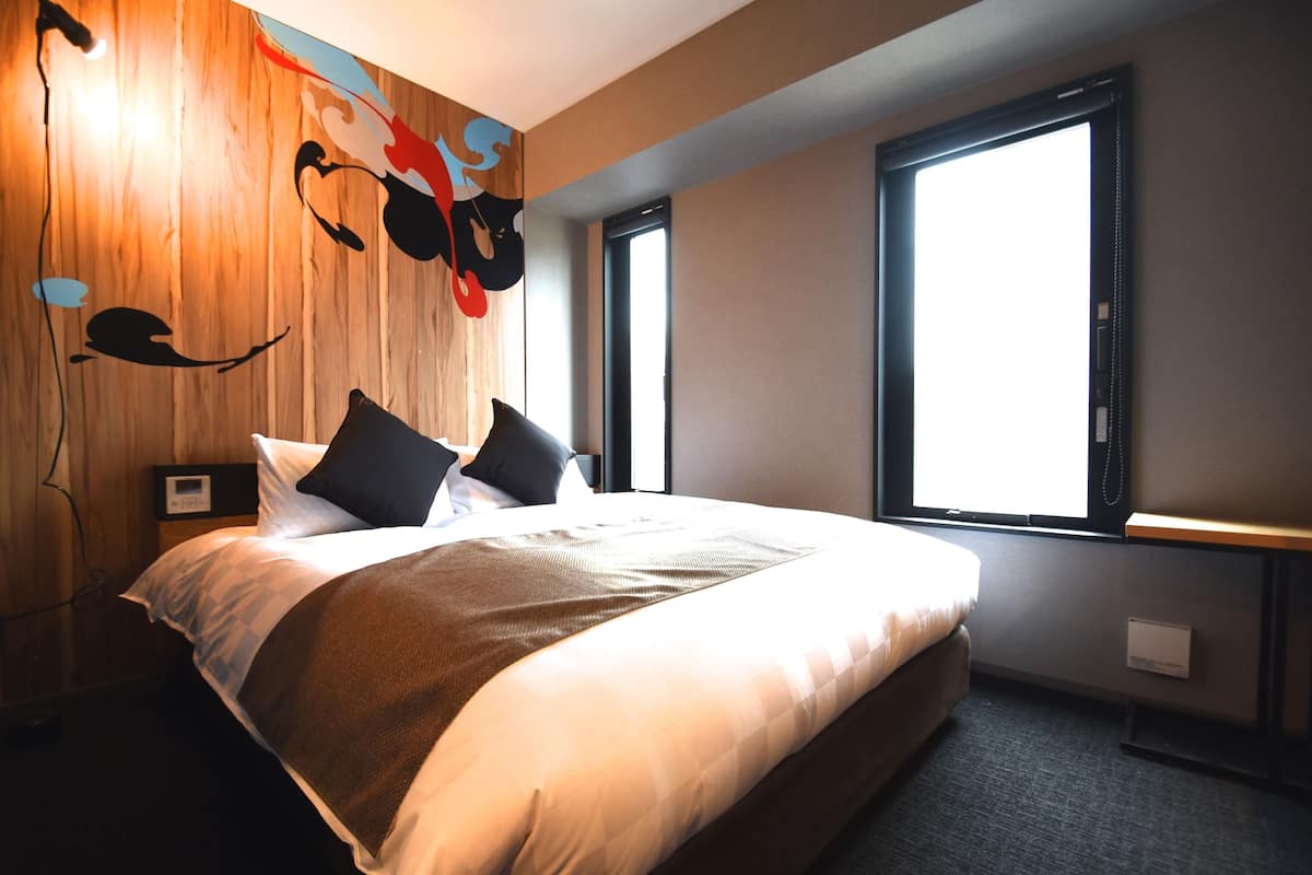 A modern hotel room with a large bed, a wooden accent wall, and a large window providing natural light.