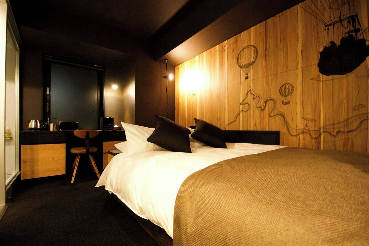 A hotel room with a wooden accent wall, a bed with white linens, a desk with a chair, and a small kitchenette.