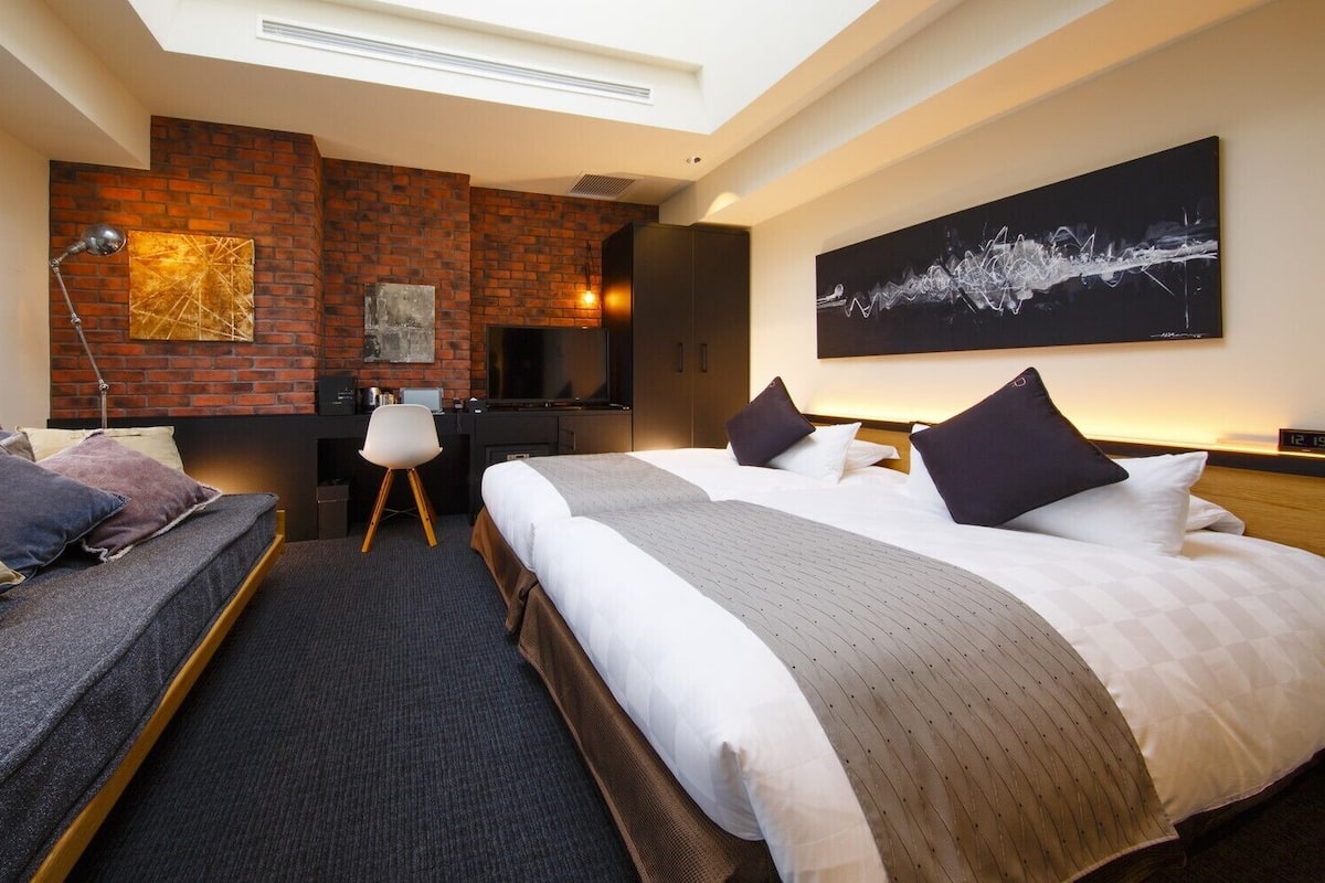 A hotel room with two beds, a desk, a chair, a brick accent wall, and artwork.