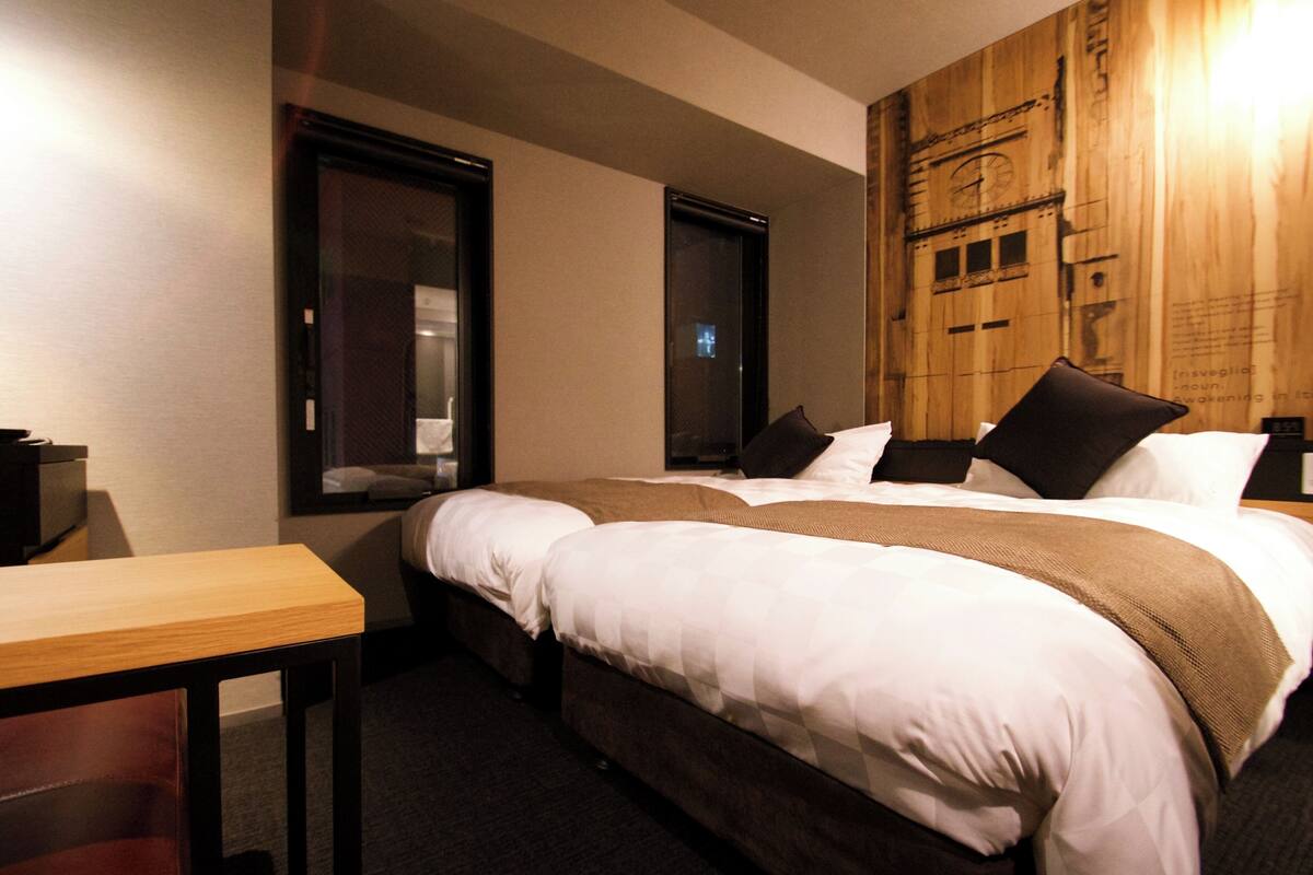A hotel room with a large bed, a wooden headboard, a small table, and a window with a view of a building.