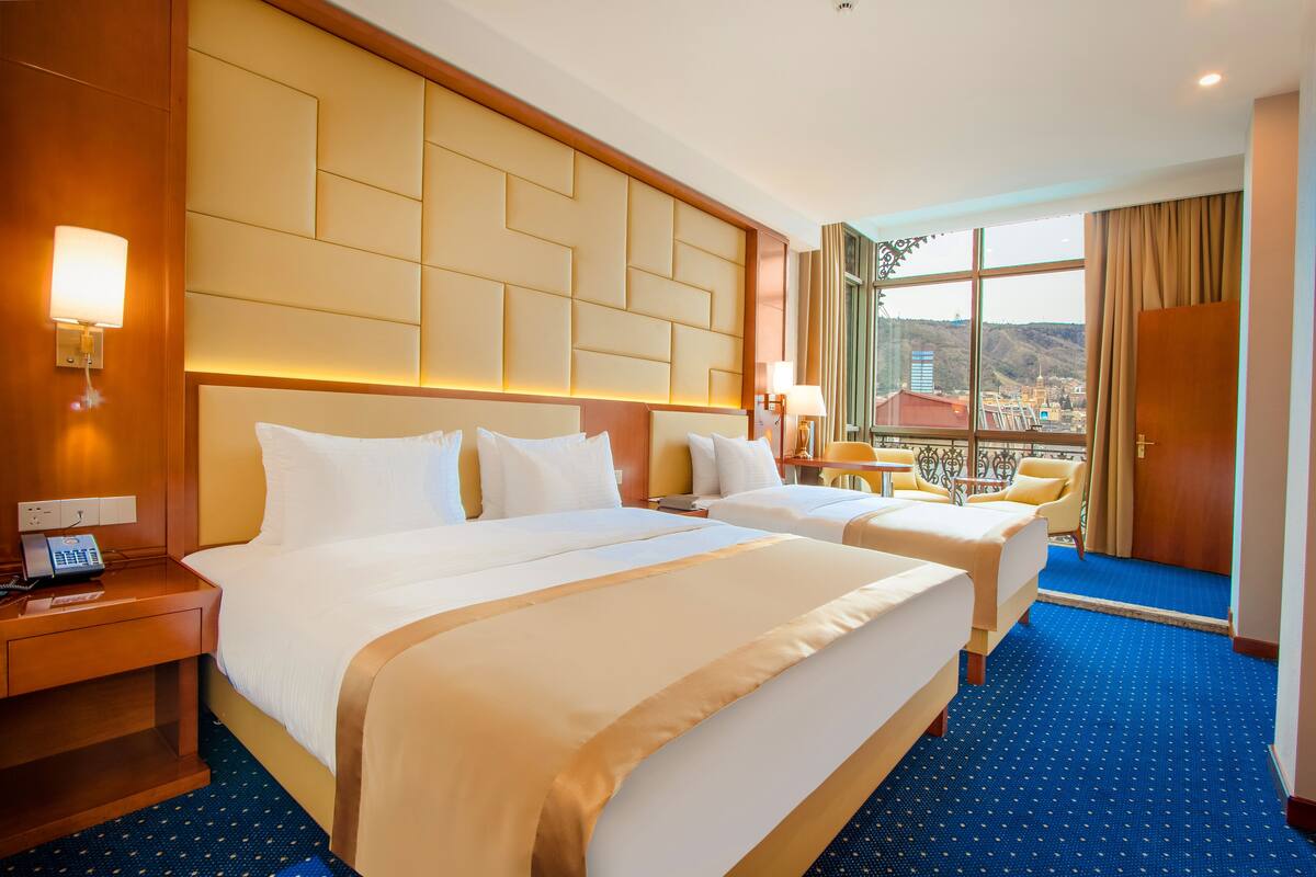 A hotel room with two beds, a large window with a city view, a wooden headboard, and a blue carpet.