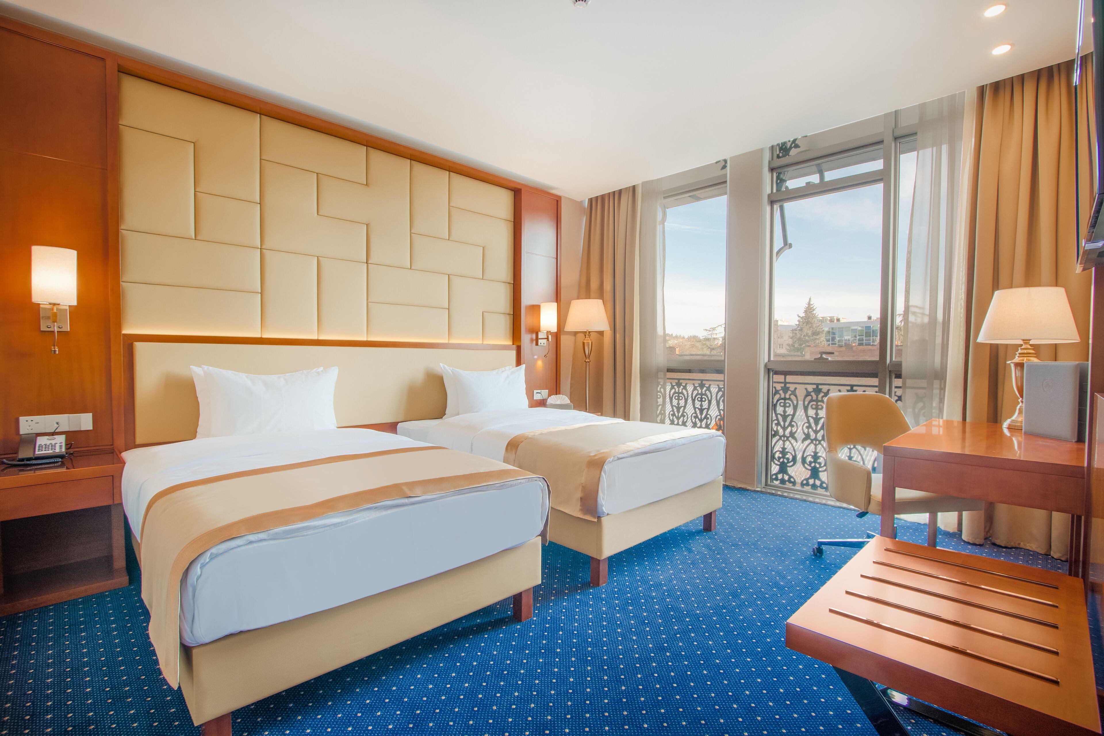 Quarto de hotel com duas camas, uma escrivaninha e uma cadeira. Há uma janela com vista para a cidade.