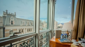A hotel room with a large window offering a view of historic buildings. The room includes a wooden desk with a lamp, water bottles, and a telephone. - New Tiflis Hotel (Tbilisi)
