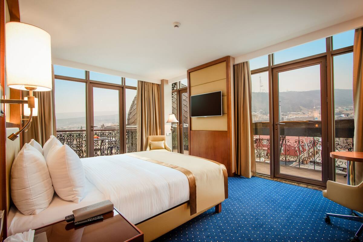 A hotel room with a large bed, a TV mounted on the wall, a chair, and a view of the city through large windows.