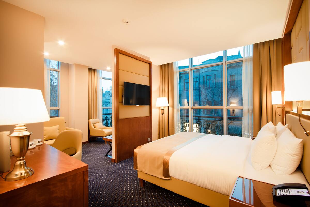 A hotel room with a large bed, a desk with a lamp, a television, and a view of a building through the window.