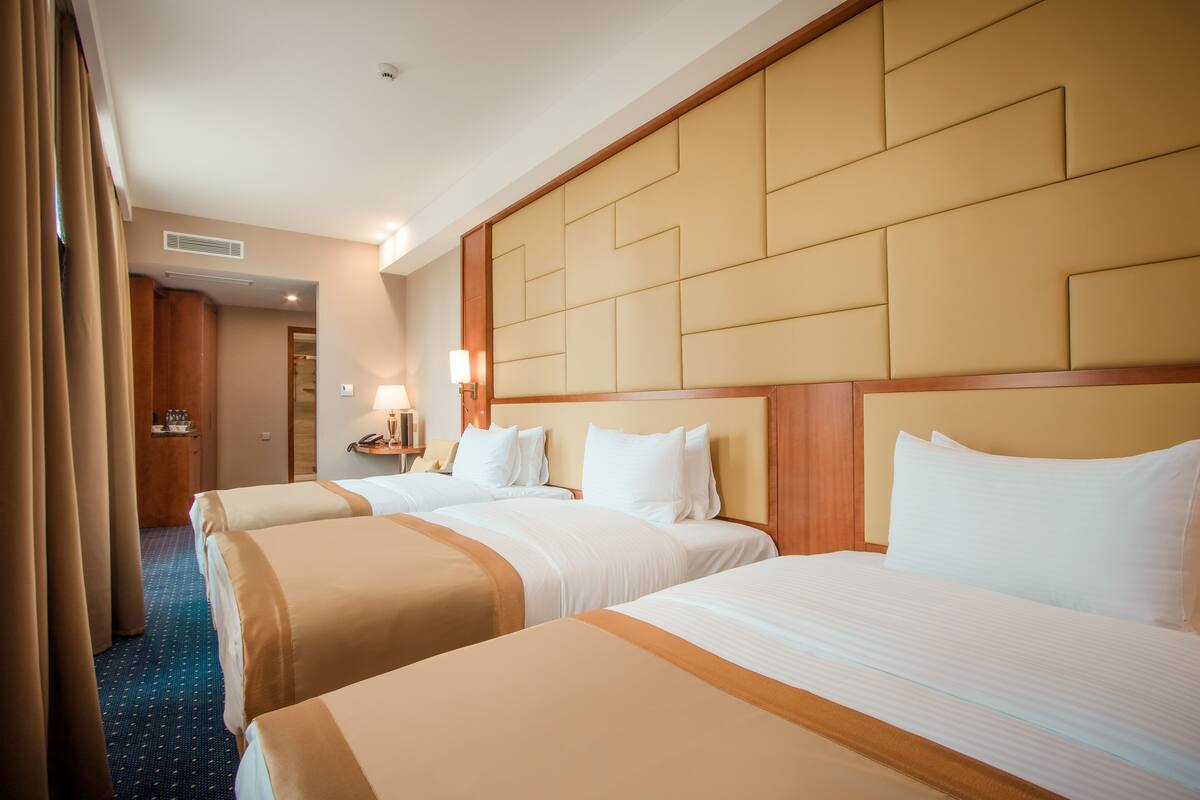 A hotel room with three beds, a wooden headboard, a blue carpet, and a nightstand with a lamp.