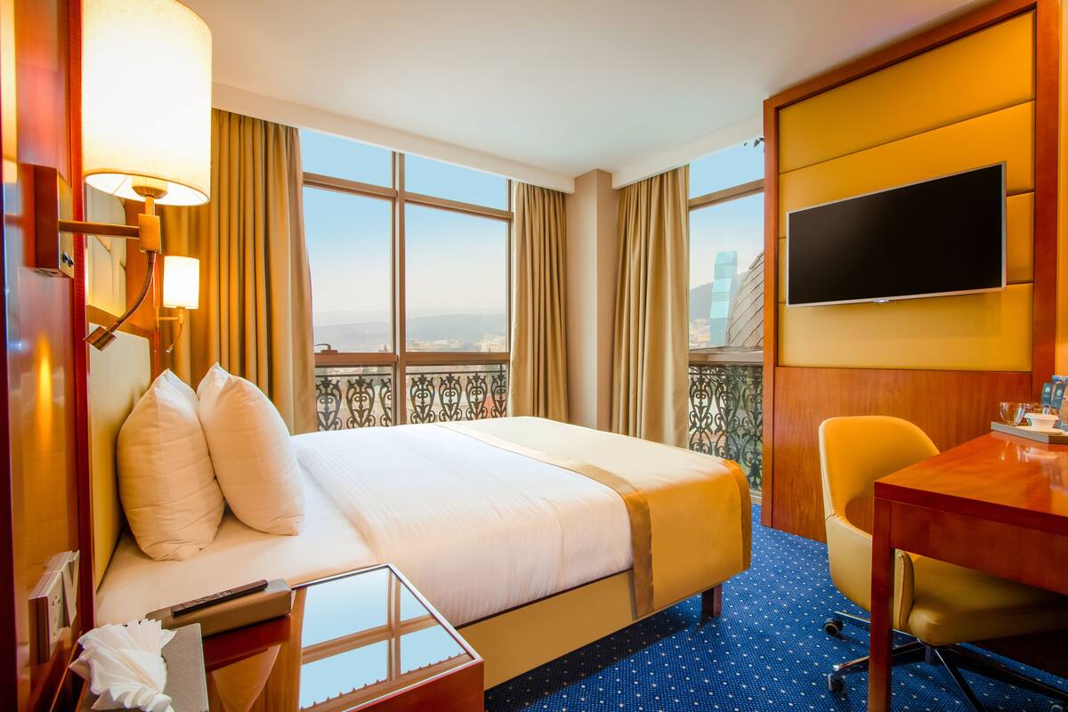 A hotel room with a bed, a desk, a chair, a TV, and a balcony with a city view.