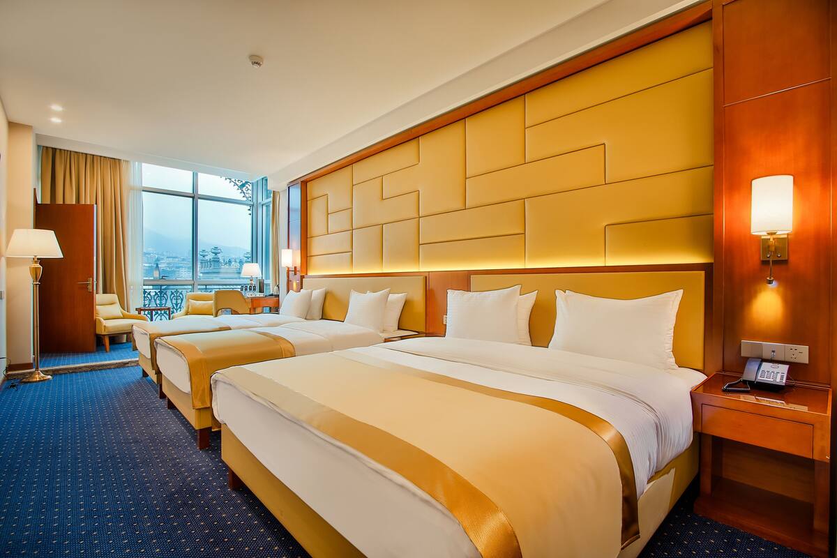 A hotel room with two beds, a large window with a city view, a wooden headboard, and a bedside table with a lamp.