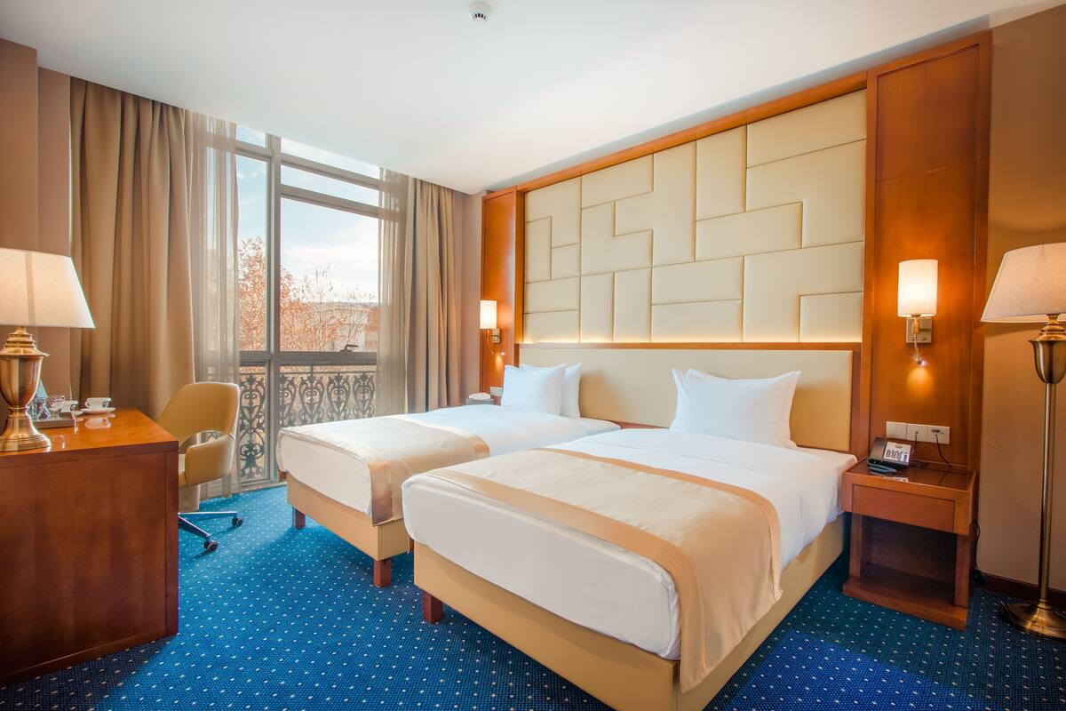 A hotel room with two beds, a desk, a chair, and a view of the outdoors.