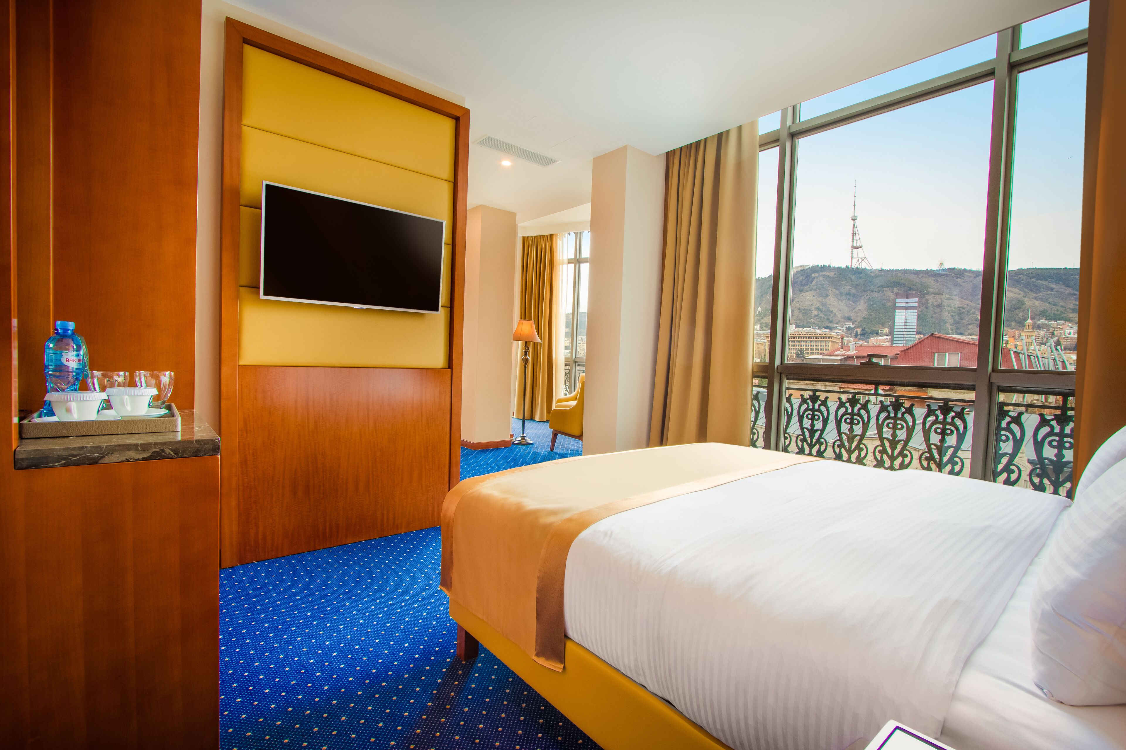 A hotel room with a bed, a flat-screen TV, a window with a view of a cityscape, and a blue carpet.