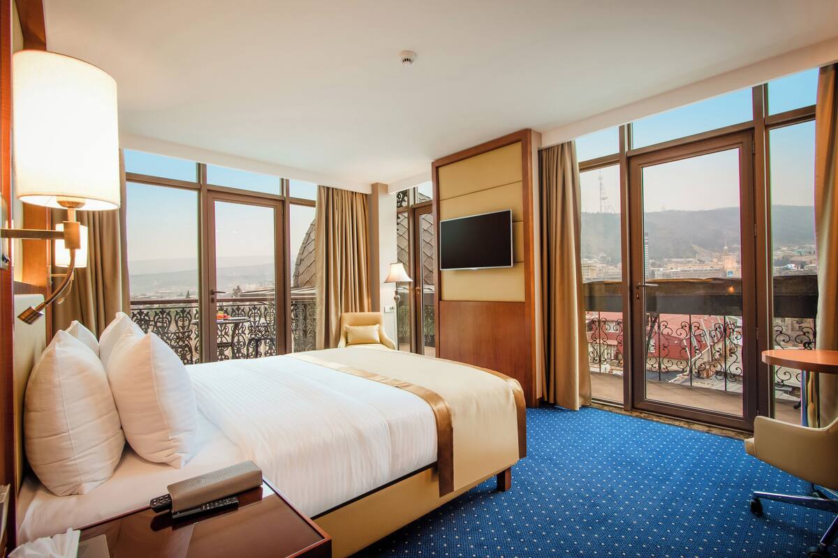 A hotel room with a large bed, a TV mounted on the wall, a chair, and a view of the city through large windows.