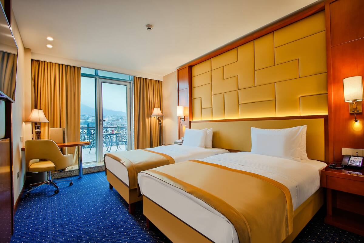 A hotel room with two beds, a desk, a chair, and a view of the city.