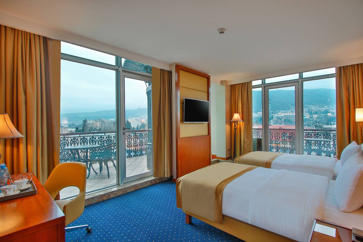 A hotel room with two beds, a desk, a chair, a TV, and large windows with a city view.