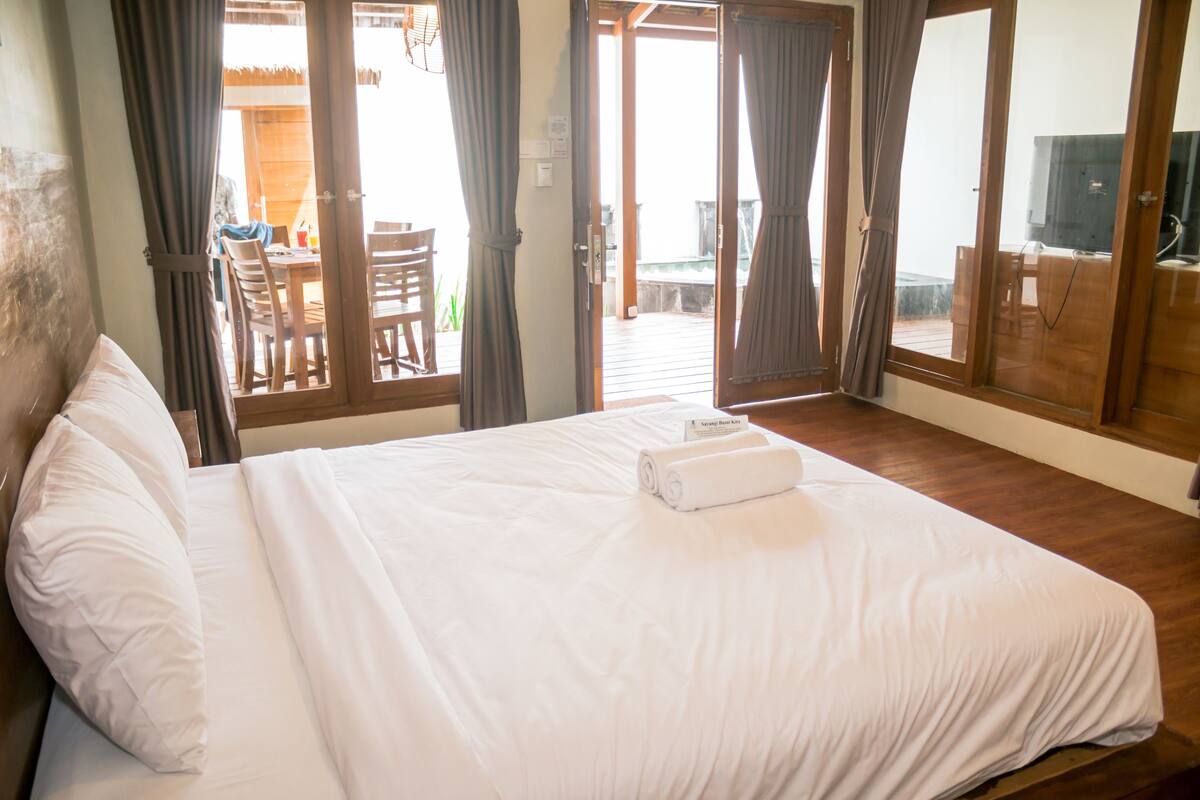 Une chambre d’hôtel avec un grand lit, un plancher en bois et une vue sur l’espace extérieur grâce à des portes vitrées.