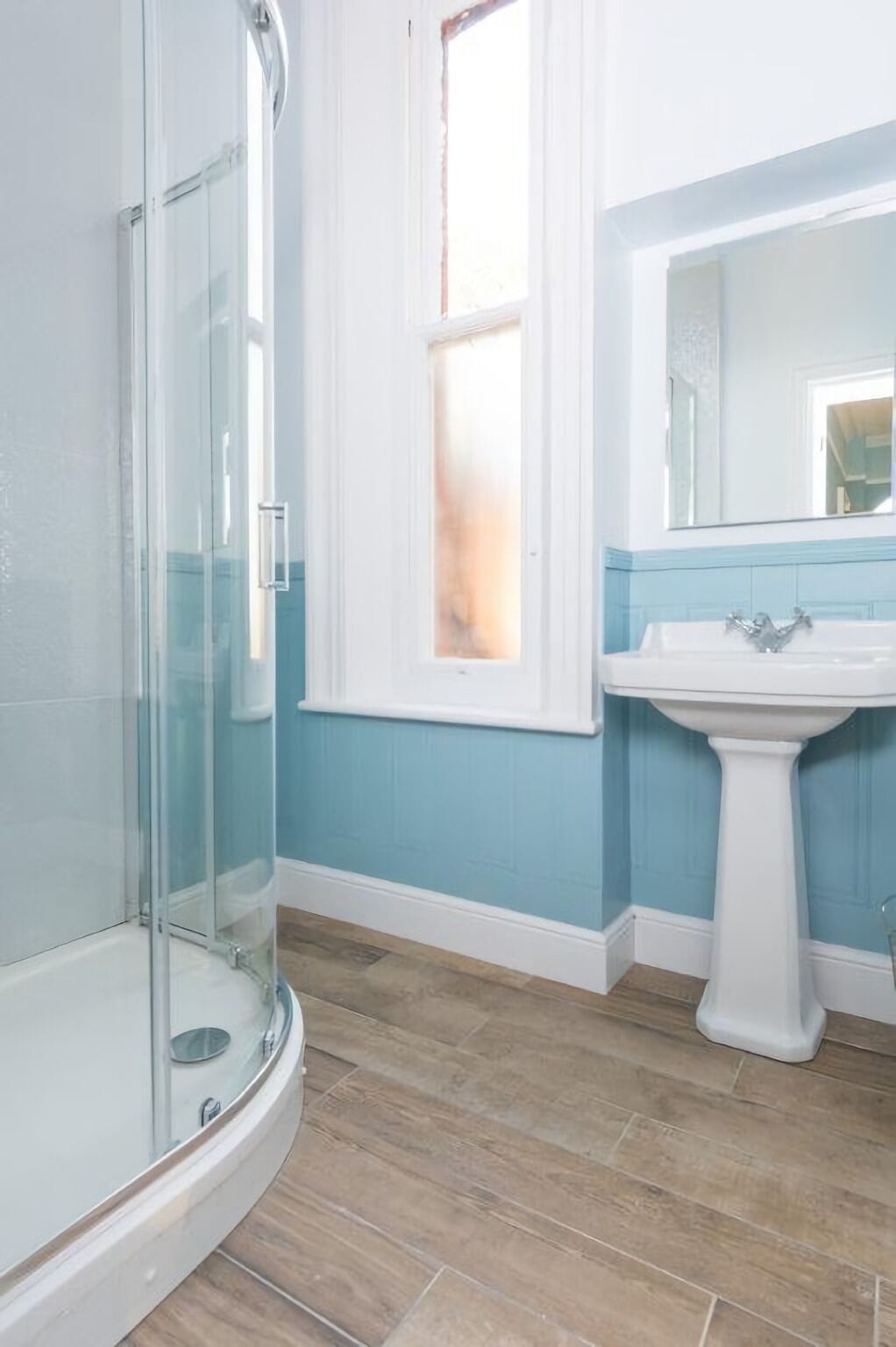 A bathroom with a glass shower enclosure, a pedestal sink, and a window with a view.