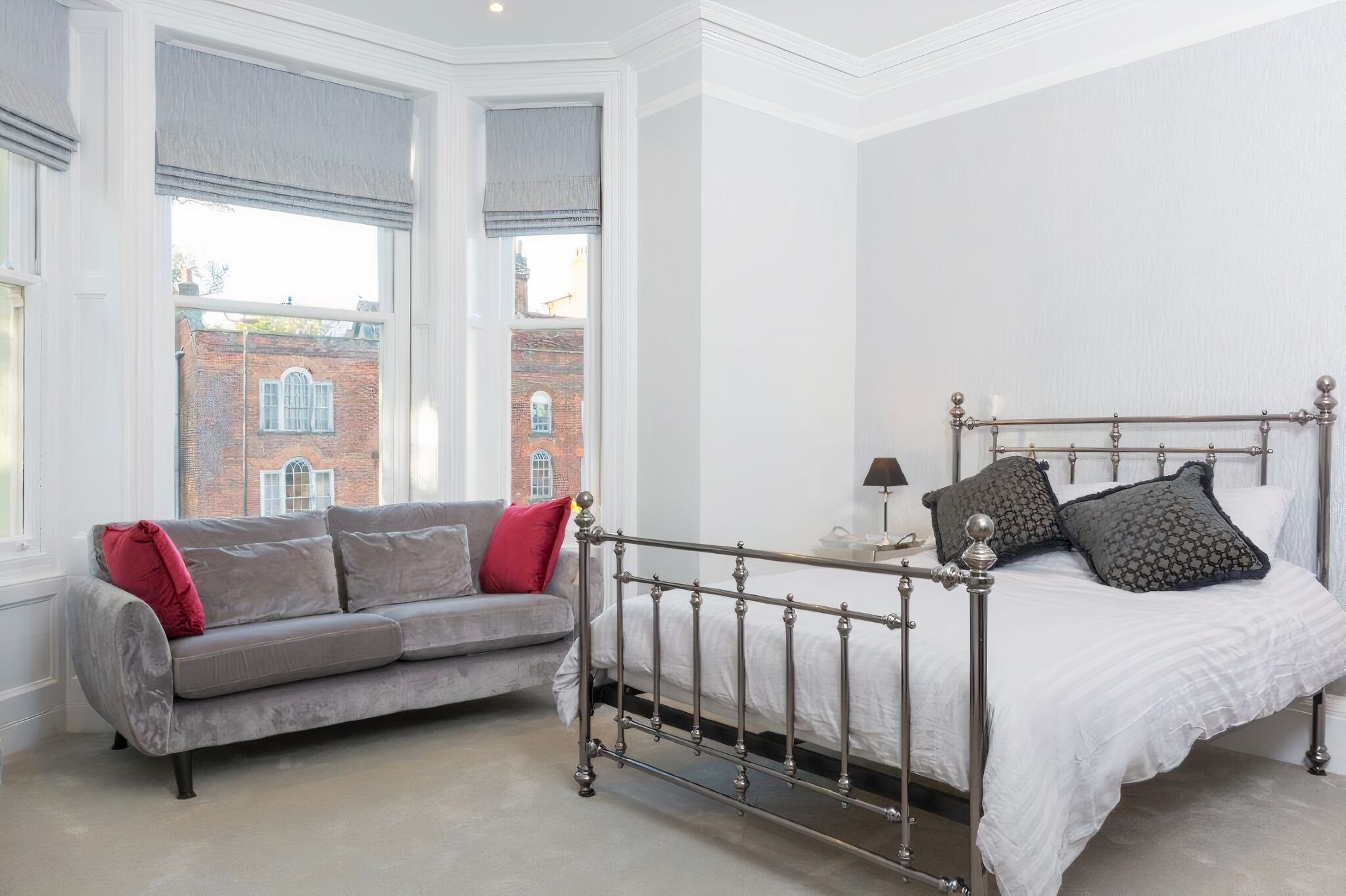 Una camera da letto con una struttura del letto in metallo, un divano grigio con cuscini rossi e vista su un edificio in mattoni attraverso ampie finestre.