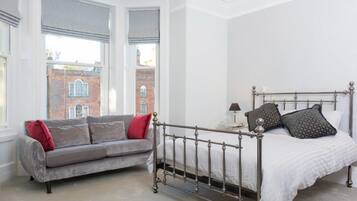 Una camera da letto con una struttura del letto in metallo, un divano grigio con cuscini rossi e vista su un edificio in mattoni attraverso ampie finestre.