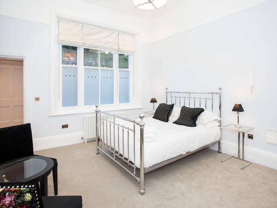 Ein Schlafzimmer mit einem Metallbett, einem Glastisch, einem Stuhl und einem Fenster mit Jalousien.