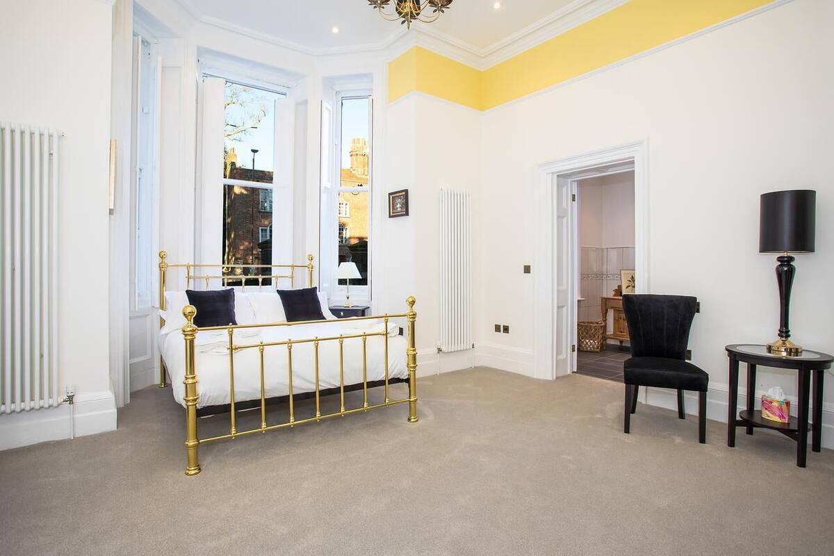 Makuuhuoneessa on kultaisella kehyksellä varustettu sänky, musta tuoli ja pienellä pöydällä oleva lamppu.