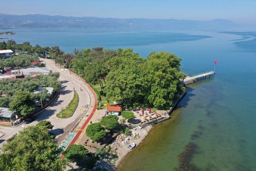 Vista aérea de una zona costera con una carretera que serpentea alrededor de un parque, un muelle que se adentra en el agua, y una vista despejada del mar y colinas distantes.
