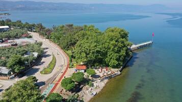 Vista aérea de una zona costera con una carretera que serpentea alrededor de un parque, un muelle que se adentra en el agua, y una vista despejada del mar y colinas distantes.