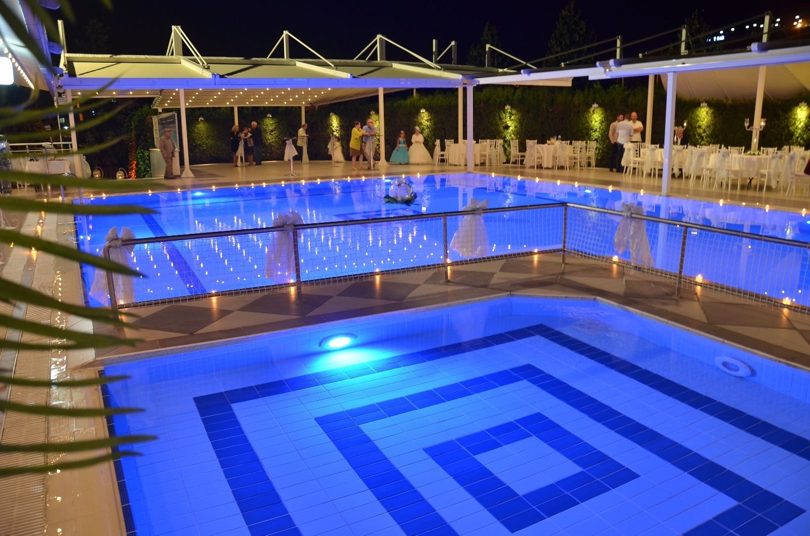 Uma grande piscina coberta com um padrão geométrico de azulejos, cercada por cadeiras e mesas brancas sob uma área coberta.
