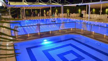 Uma grande piscina coberta com um padrão geométrico de azulejos, cercada por cadeiras e mesas brancas sob uma área coberta.