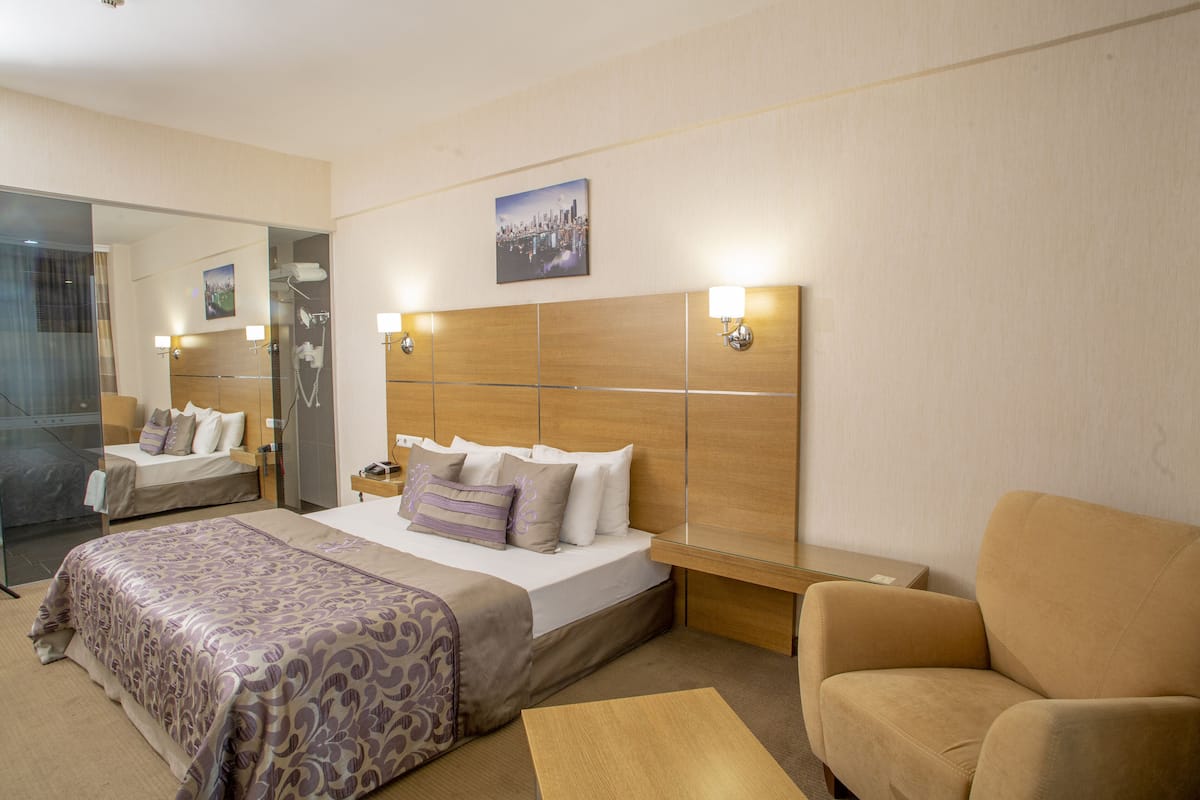 A hotel room with a large bed, a sofa, a bedside table, a mirror, and a cityscape painting on the wall.