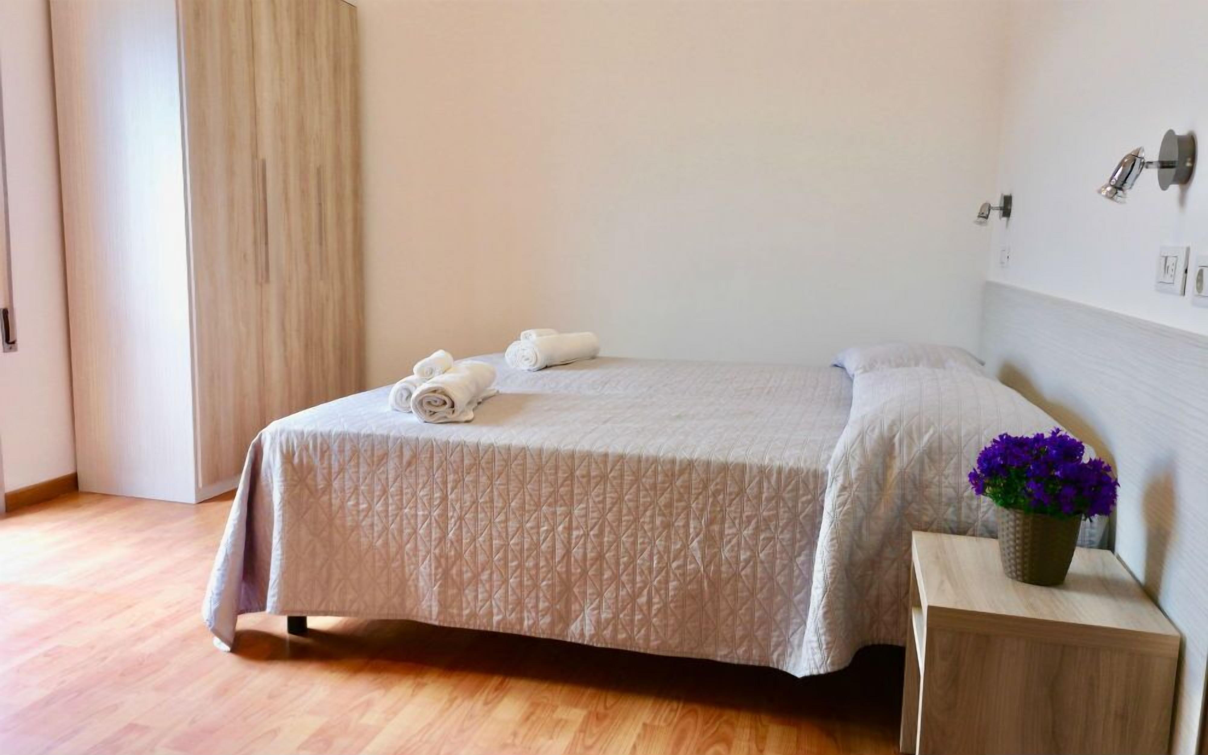A neatly made bed with a quilted cover, a bedside table with a vase of flowers, and a wall-mounted lamp.
