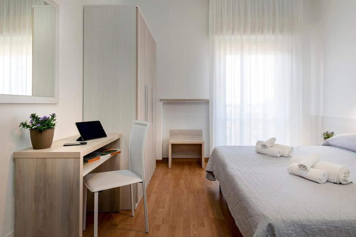 A modern hotel room with a wooden desk, a chair, a bed with neatly folded towels, and a small side table.