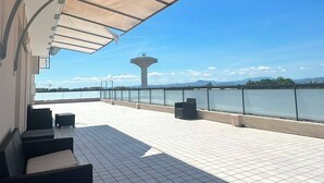 An outdoor terrace with tiled flooring, a covered seating area, and a view of a body of water and distant landscape.