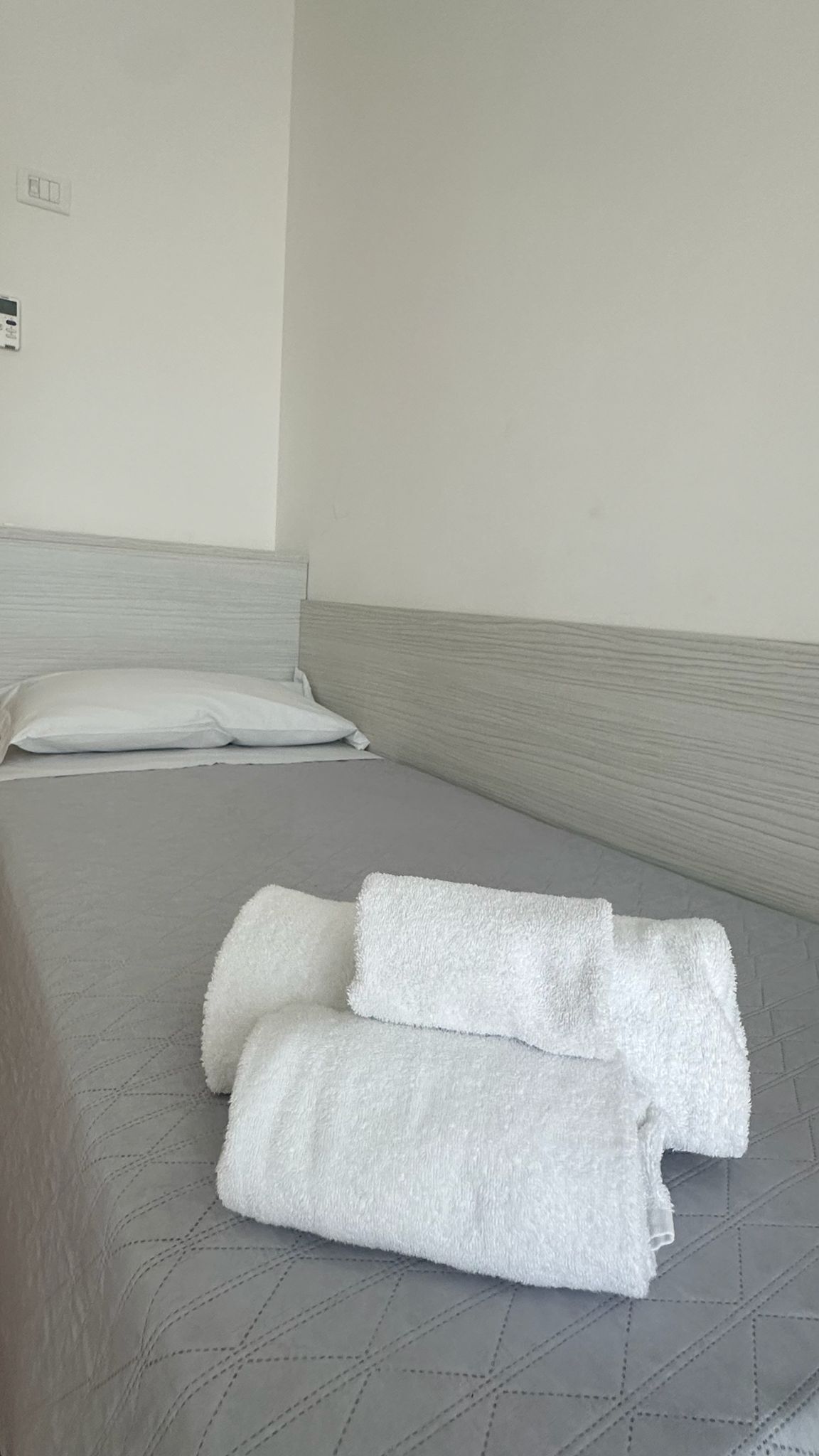 A bed with a grey quilted mattress, two rolled white towels, and a white pillow.