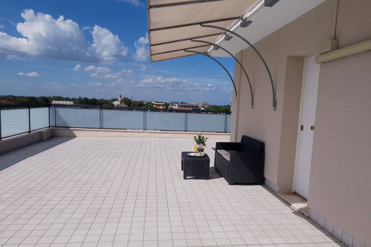 A rooftop terrace with a tiled floor, a covered seating area, and a view of the sea and buildings in the distance.