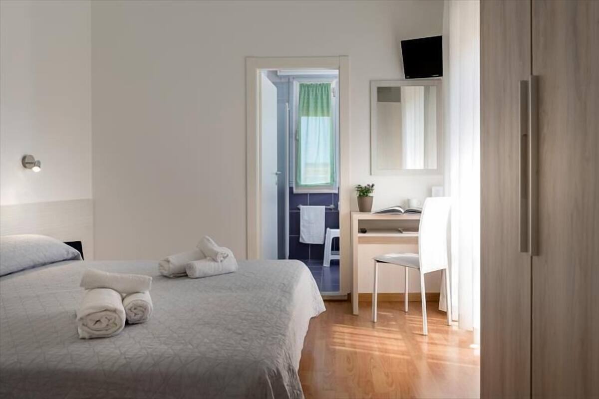 A hotel room with a bed, a desk with a chair, a television, and a bathroom visible through an open door.