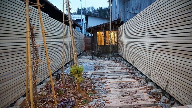 A narrow wooden pathway flanked by bamboo structures and a small plant in a garden area.