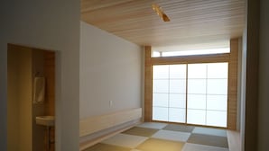 A room with tatami flooring, a low wooden shelf, a window with a grid pattern, and a wooden ceiling.